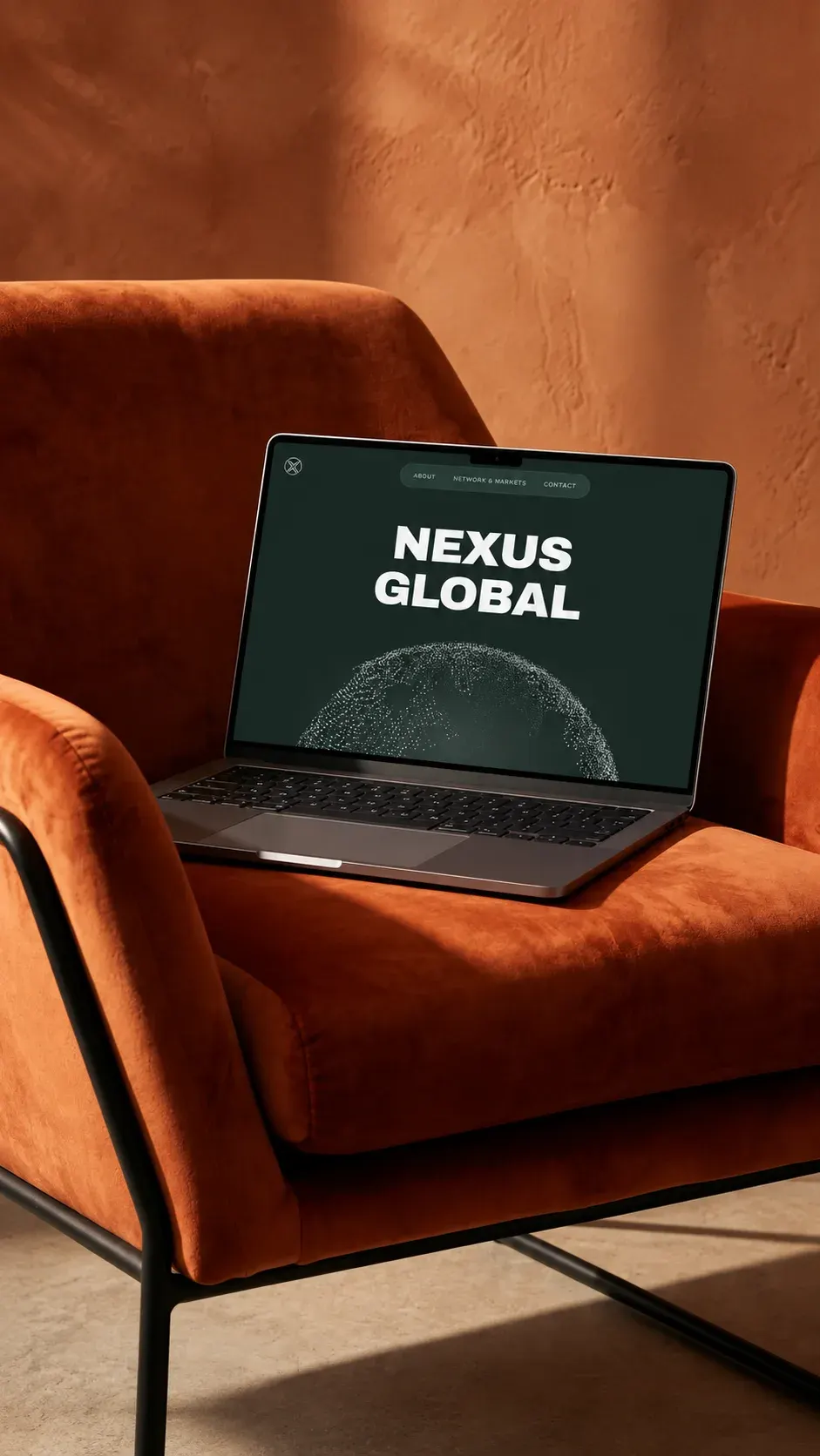 Laptop displaying the Nexus Global website, centered on a rust-orange armchair in a warm, sunlit interior with textured terracotta walls.