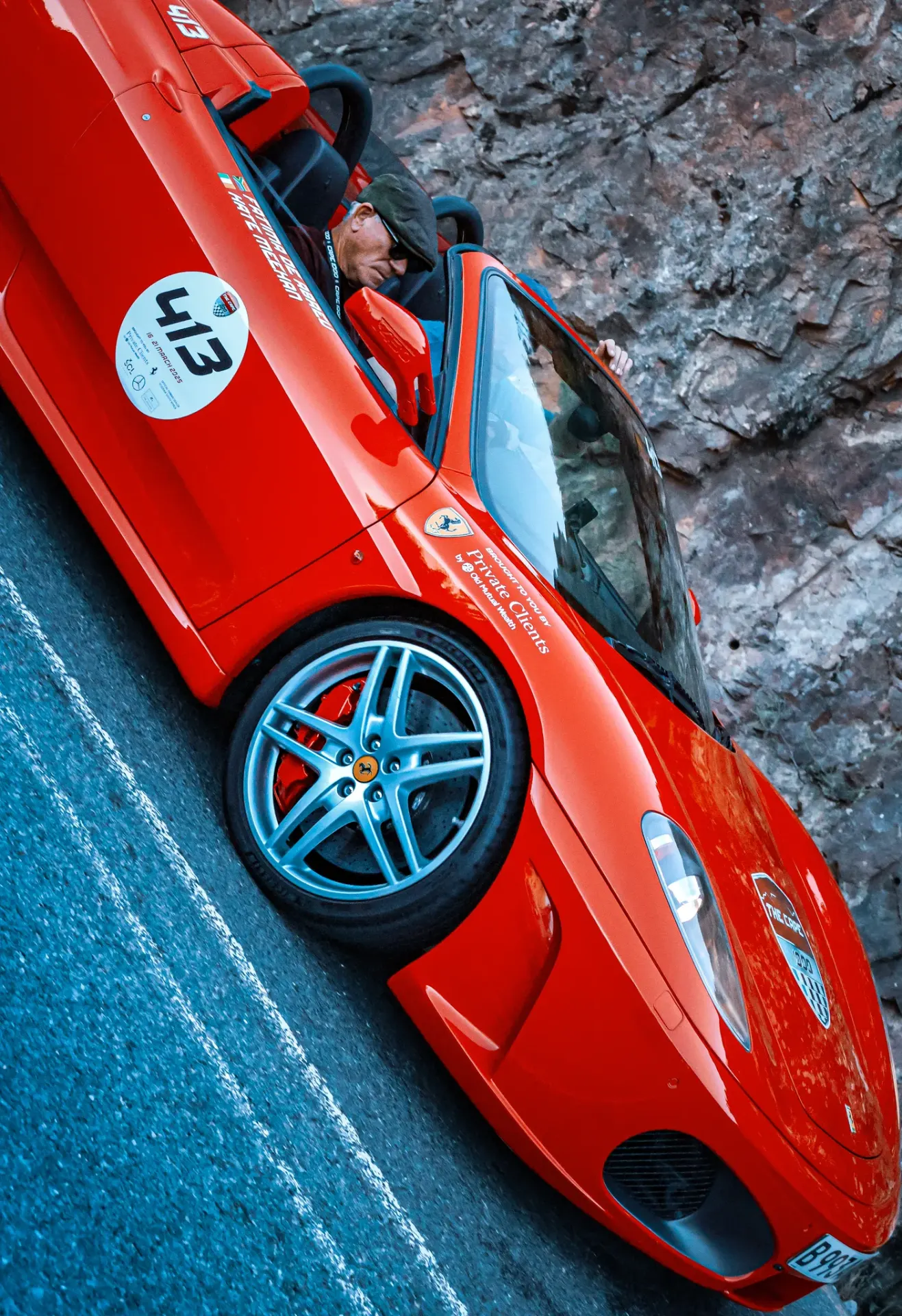 Red Ferrari sports car taking part in the Cape 1000, photographed beside a rocky mountain road as the driver steps into the vehicle, with event decals and racing number 43 visible on the bodywork.